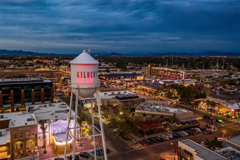 A large white water tower with black text. A water tower with a red light. A building with lights on. A blue sky with clouds. A rooftop of a building. A red roof with lights. A building with a balcony. A city at night with mountains in the background.